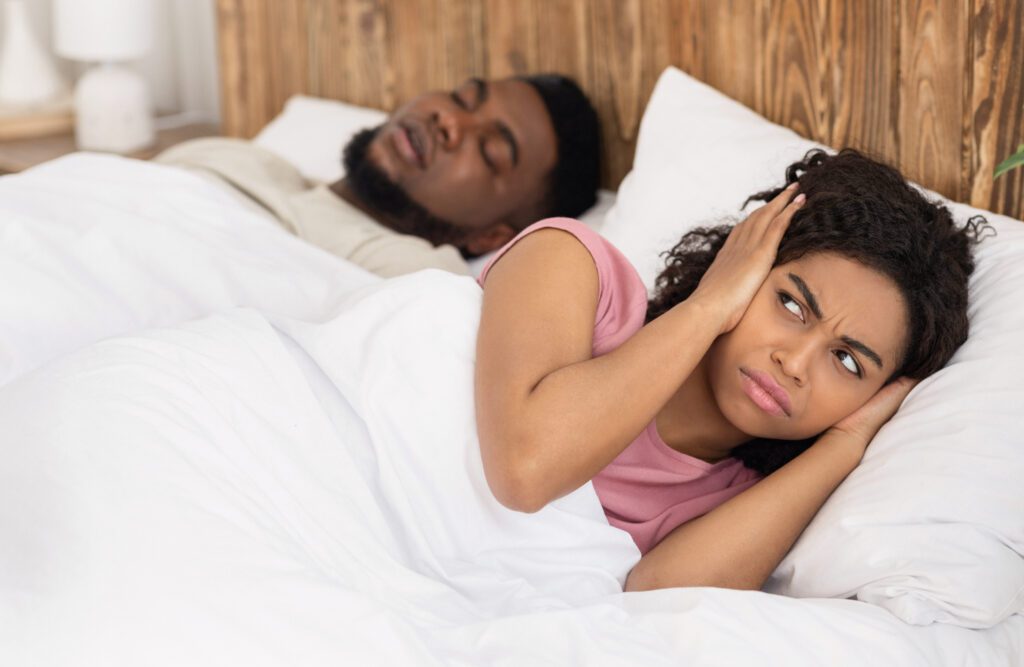 Annoyed african american woman covering her ears with palms while her boyfriend snoring at night, cannot sleep