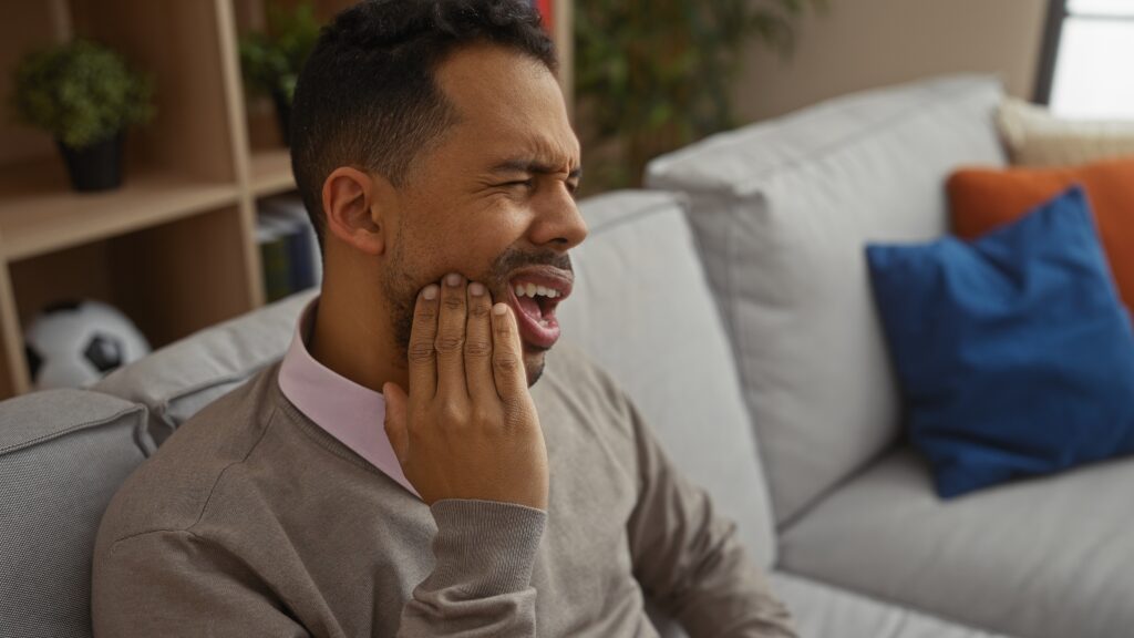 Young man experiencing pain holding his jaw while sitting on a couch in a cozy home living room showcasing discomfort in a modern apartment setting.