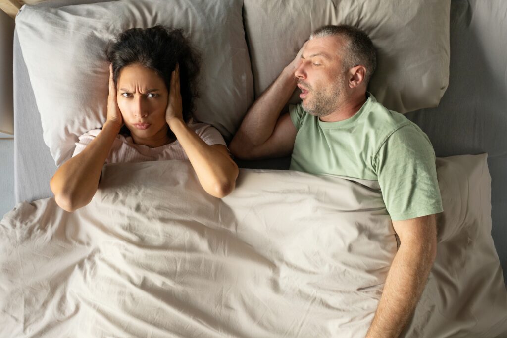 man snoring in bed while his partner covers her ears