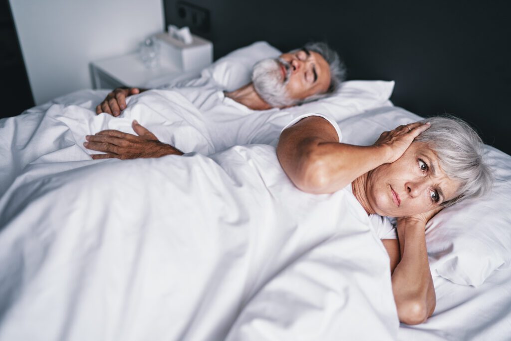 older couple in bed, wife covering her ears as husband snores